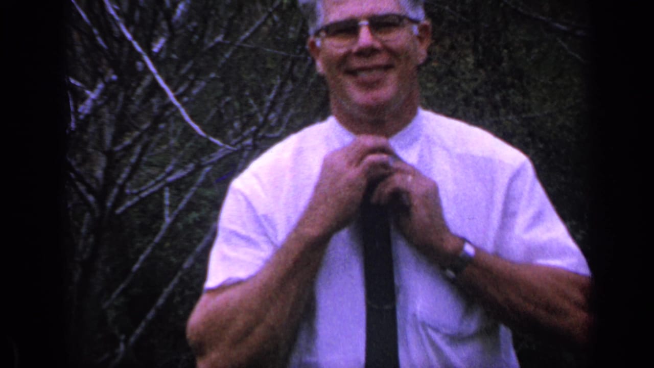 Man in glasses adjusts a dark tie outdoors against bare branches.
