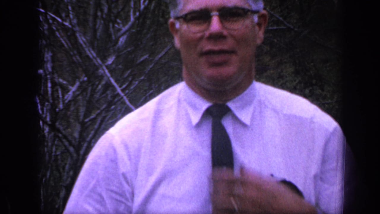 Man in white shirt and dark tie stands closer to the camera outdoors.