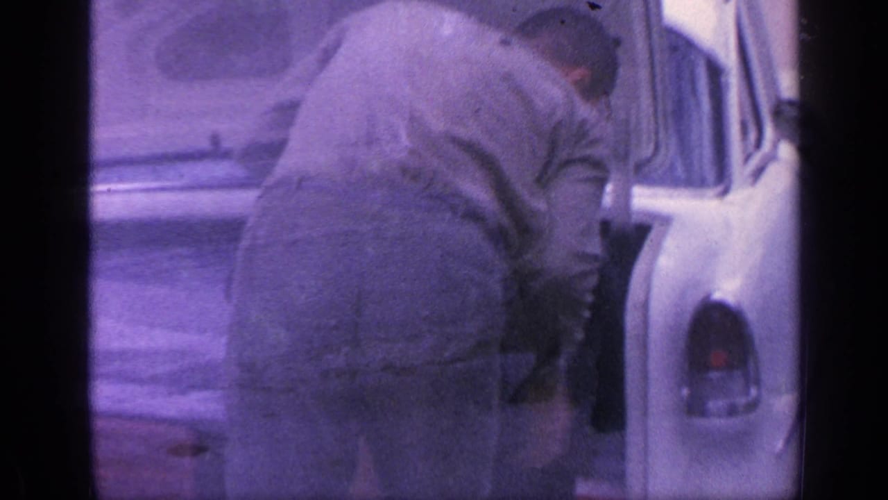 Man leaning into the open trunk of a white car, with another vehicle area partly visible.