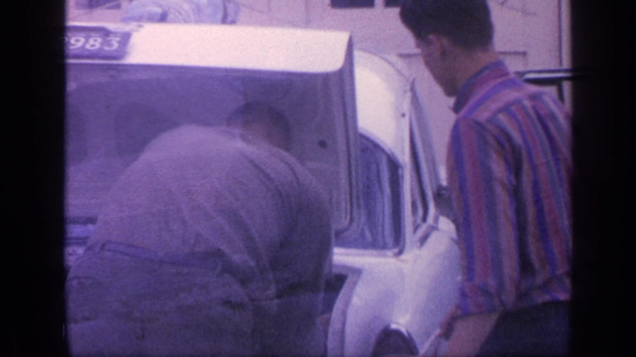 Two men beside the rear of a white car with its trunk open and license plate visible.