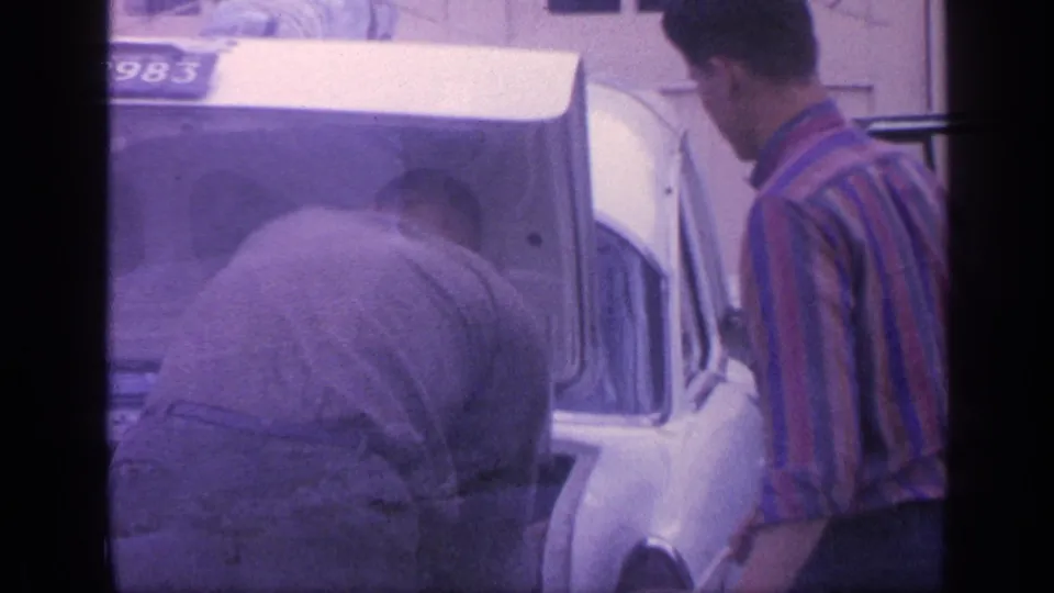 1968 Florida frame with one man bent into the trunk of a white car while a man in a striped shirt stands nearby.