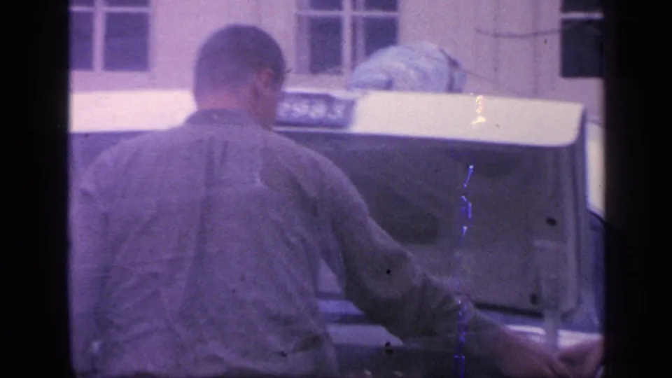 1968 Florida home-movie frame showing the back of a man at the open trunk of a white car, seen through soft purple-shifted film.