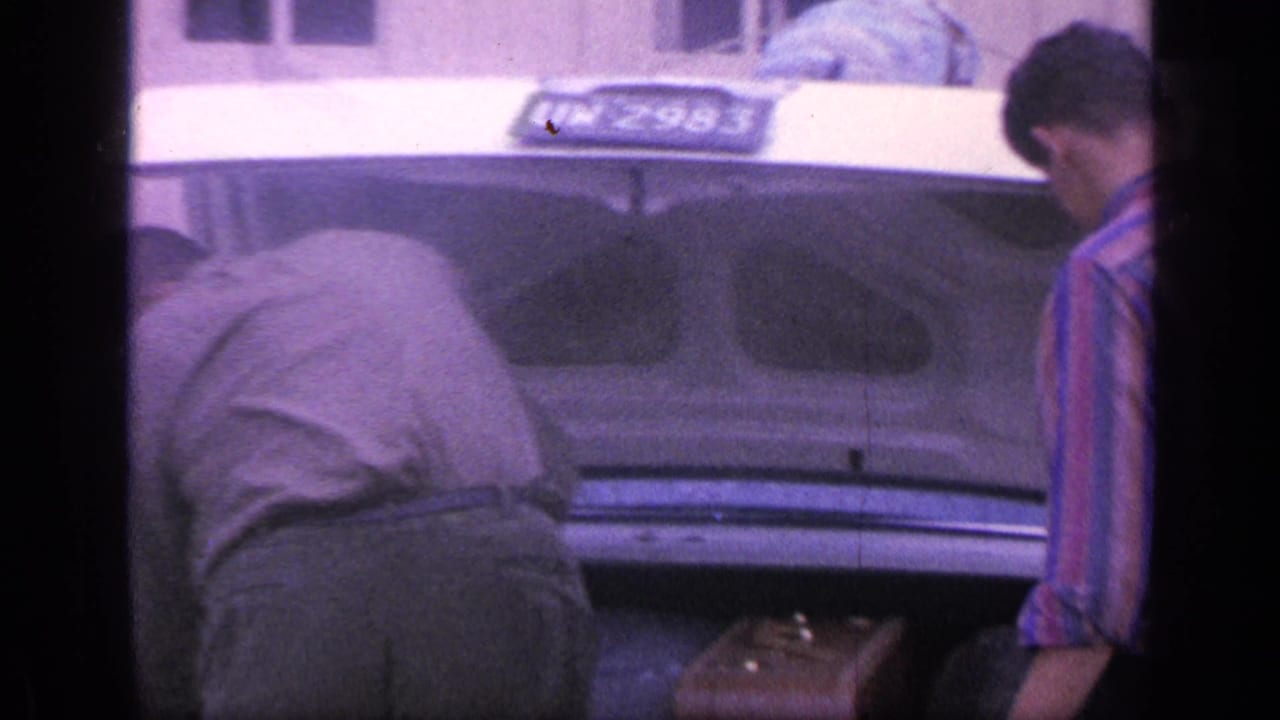 Two men stand at the open trunk of a car with luggage partly visible.