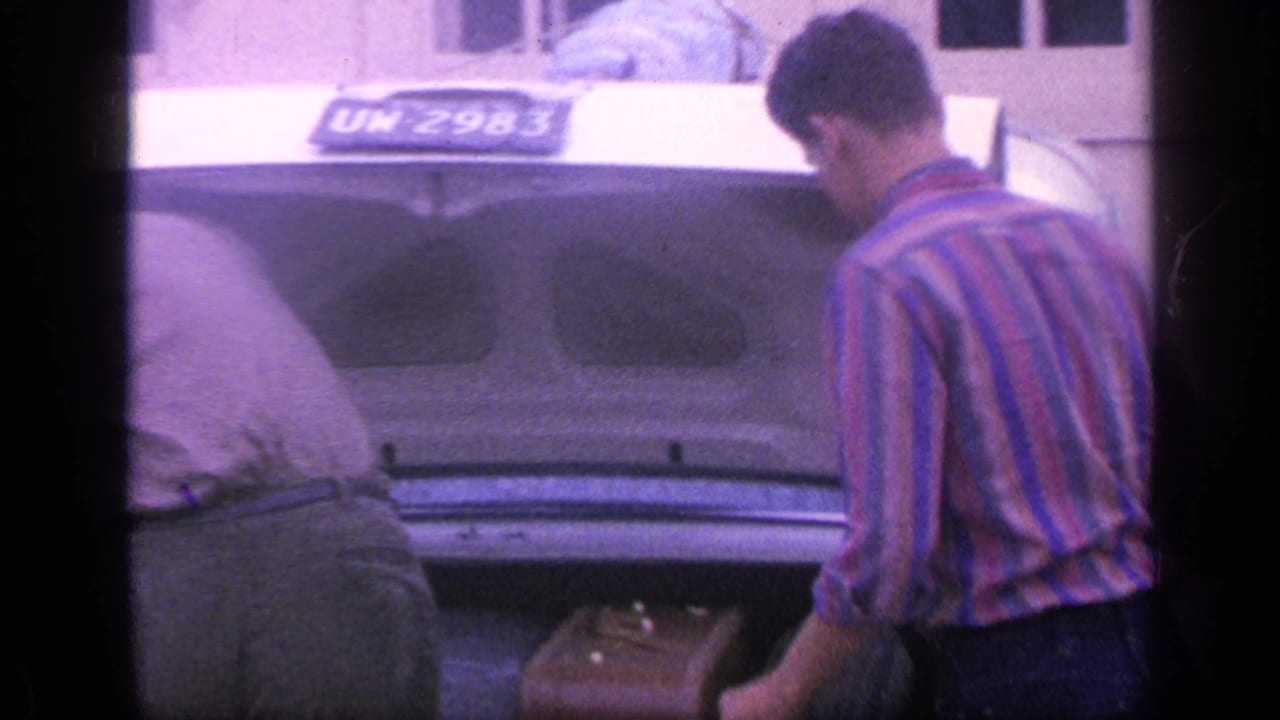 A man in a striped shirt stands beside a car trunk while another bends inside it.