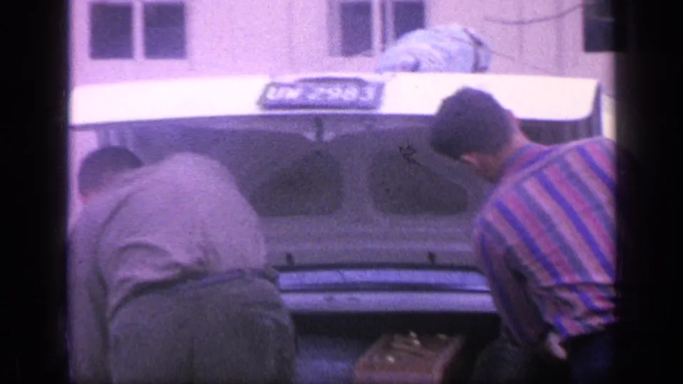 Two adult men lean over the open trunk, continuing to arrange luggage in the 1968 Florida home-movie scene.