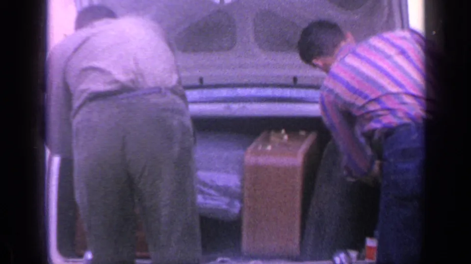 At the end of the clip, two men bend into the open trunk while an upright suitcase sits next to the rear bumper.