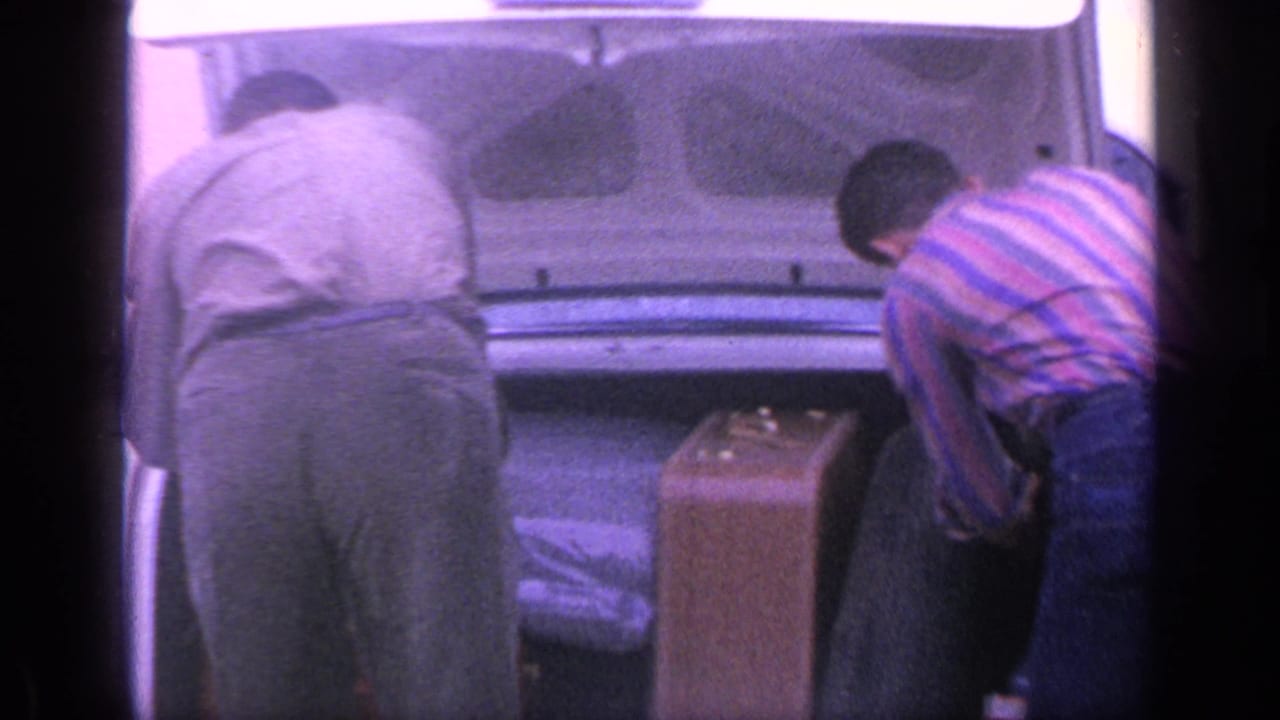 Two men bend over the open trunk of a white sedan with a brown suitcase nearby.