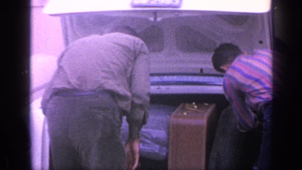 A man in a light shirt reaches into an open car trunk while another man bends nearby.