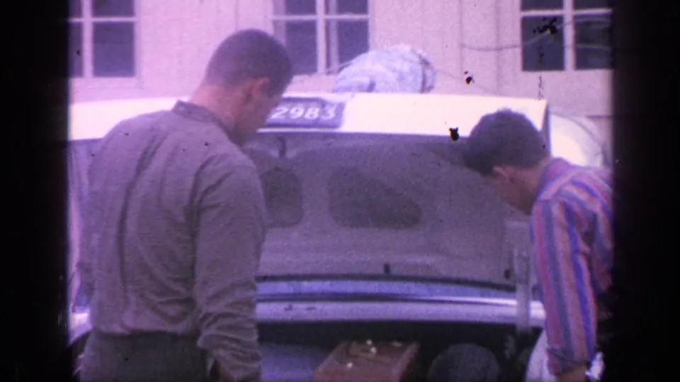 The white sedan's open trunk and visible plate are centered as two men look down toward luggage near the rear of the car.
