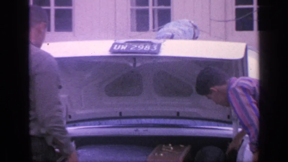 At the end of the clip, two adult men stand near the open trunk of a white sedan, with a brown suitcase visible inside or beside it.