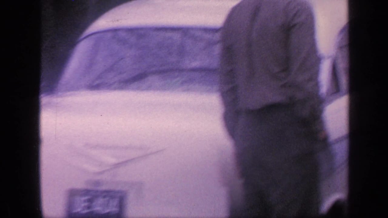 Man stands near the rear of a white car seen through a soft film frame.