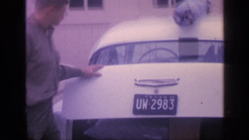 The man leans at the back of the white car while a blue Texas license plate and chrome trim are visible.