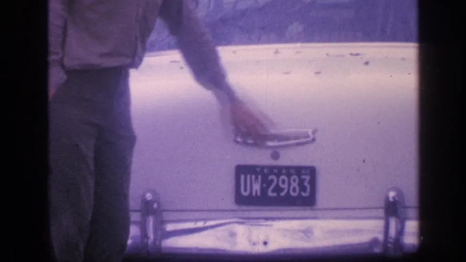 A man reaches across the closed trunk of the white classic car, with the Texas plate and chrome bumper visible.