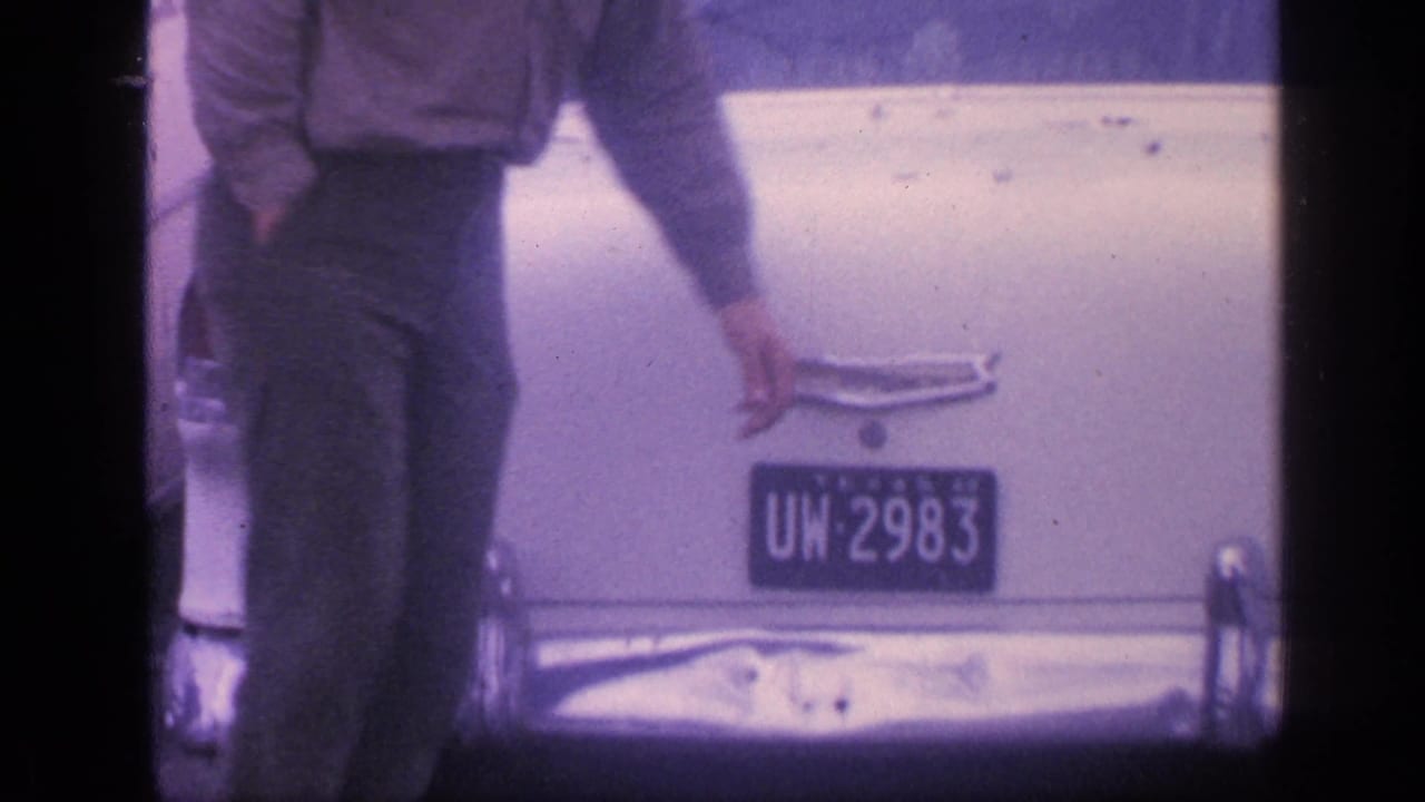 Man standing by the rear of a white sedan with a Texas license plate.