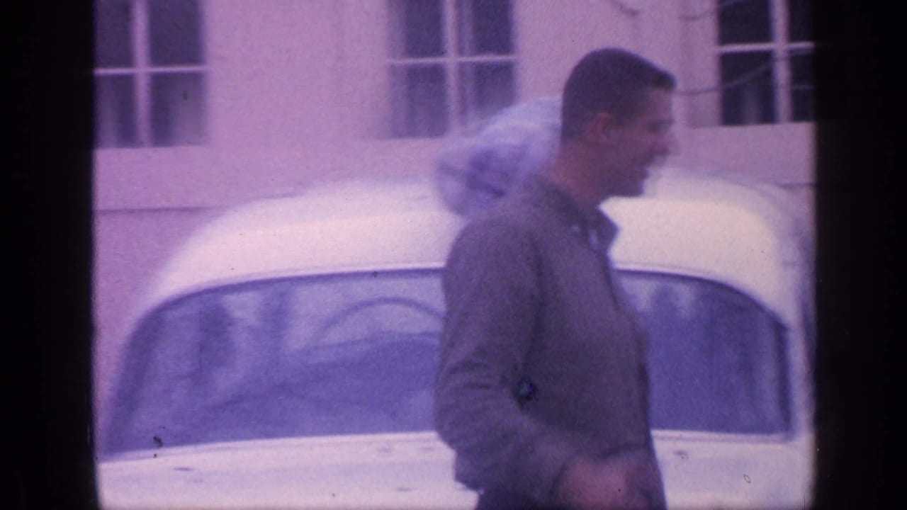 Man walking past a white sedan in front of a light building.
