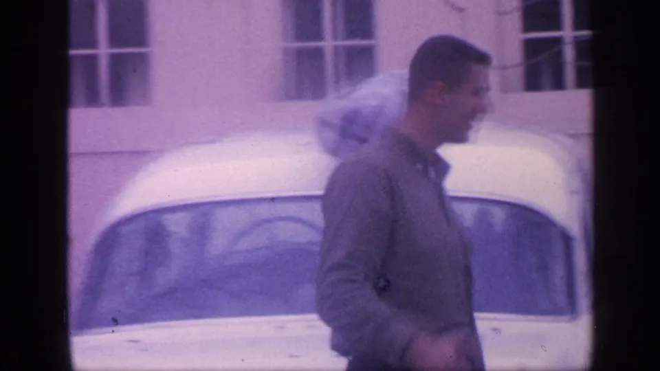 1968 Florida frame of a man walking beside a white sedan parked near a light building with windows.
