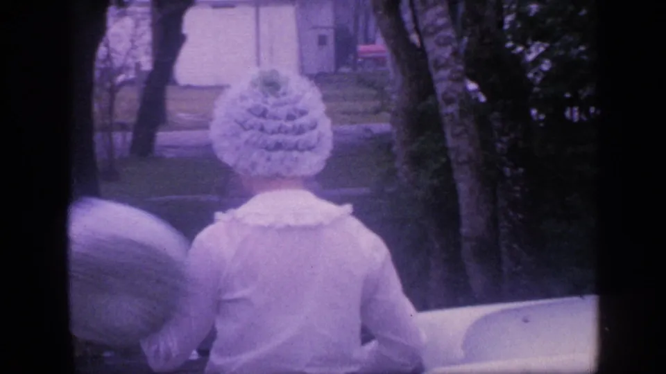 1968 Florida home-movie frame showing a woman in a white ruffled hat near the white sedan and roadside shrubs.