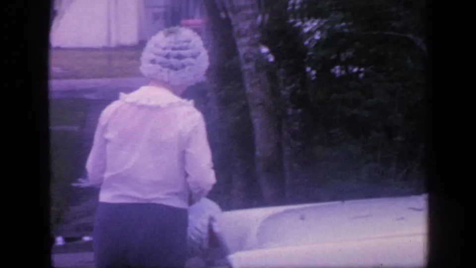 1968 Florida frame with the woman near the white sedan and the man partly visible beside shrubs and a light building.
