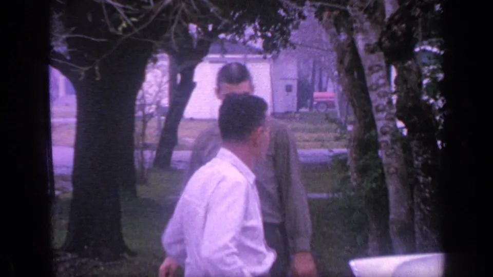 Two adults stand near a white car in a Florida residential yard, with trees, lawn, sidewalk, and a small house in the background.