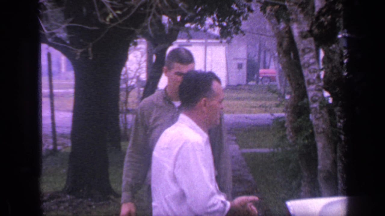 Two adult men walk near trees with part of a light-colored vehicle at the lower right.