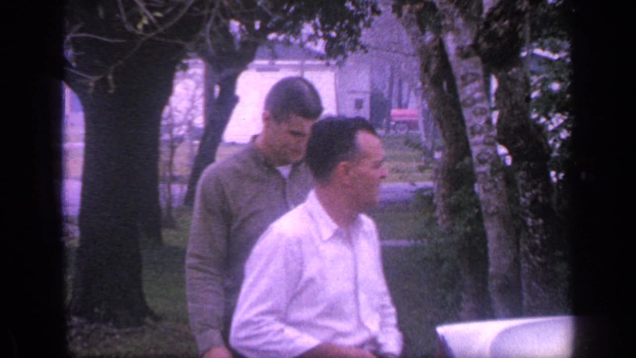 An older man in a white shirt and a younger man in a green sweater stand beside a vehicle.