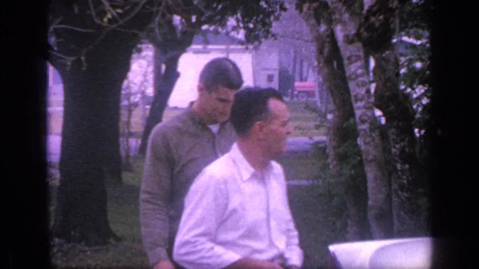 The men pause near a light-colored vehicle in a Florida yard, with trees and a pale building visible behind them.