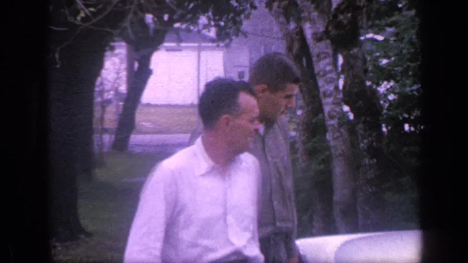 In the soft 8mm frame, the two men look downward beside the vehicle while standing in a residential Florida yard.
