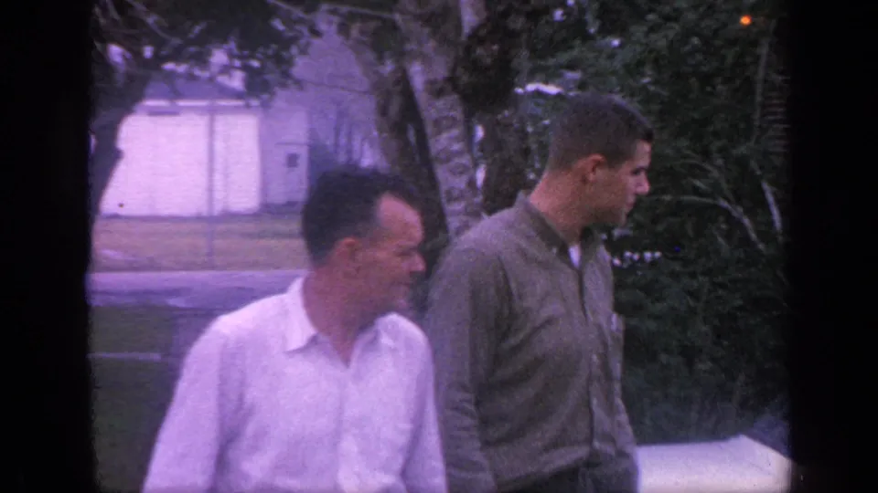 The men move rightward past the vehicle, framed by dark trees and faded 8mm color in the Florida home-movie scene.