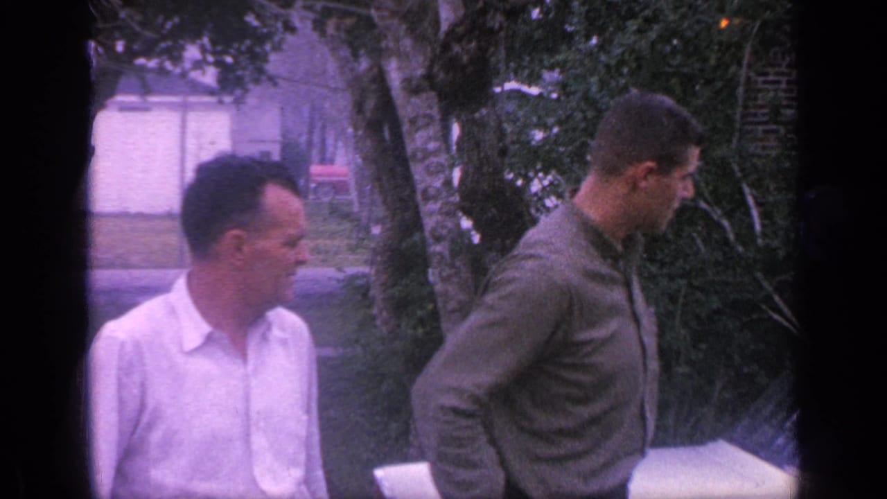 In 1968 Florida home-movie footage, two adult men stand in a yard near trees, with the older man in white facing the younger man in green.