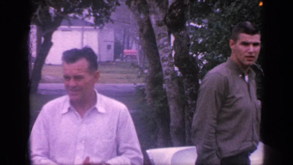 The older man smiles toward the camera or someone nearby while the younger man in green stands near trees in the 1968 Florida yard scene.