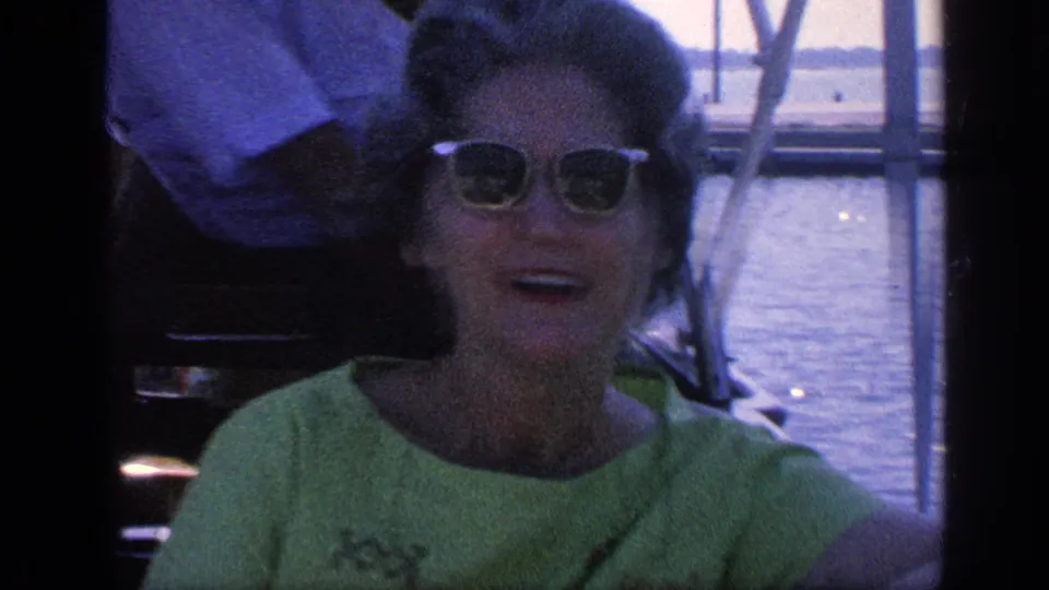1968 Florida home-movie view of a woman in a green dress and sunglasses smiling on a boat with water in the background.