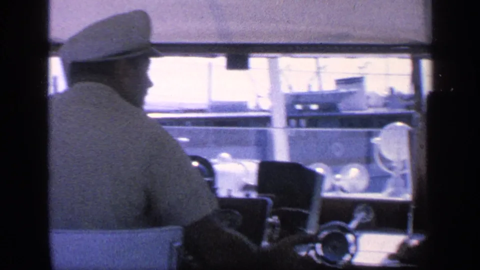 Man in a white cap, likely at the boat helm, seen in profile with water, railings, and dock structures in the background.