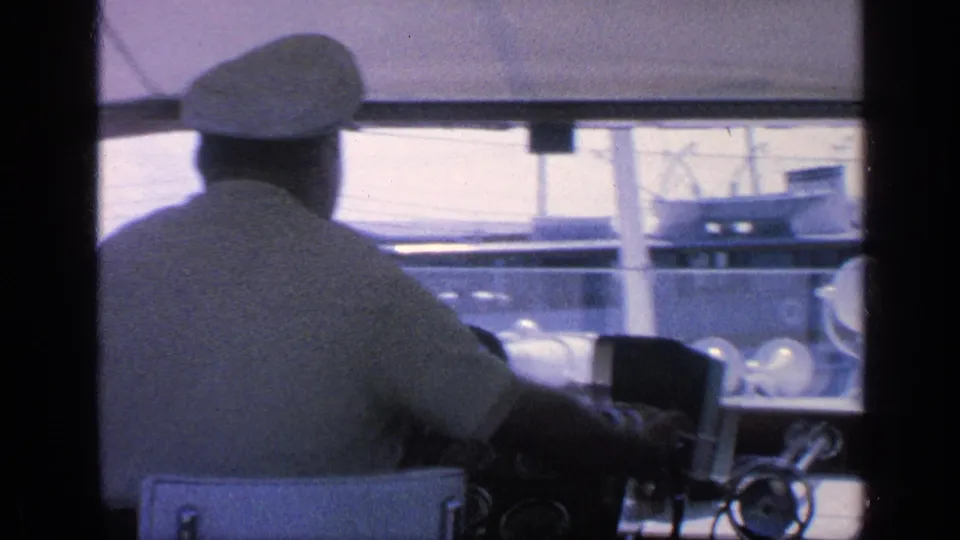 1968 Florida home-movie frame showing a captain in a white cap at the yacht helm with marina structures visible ahead.