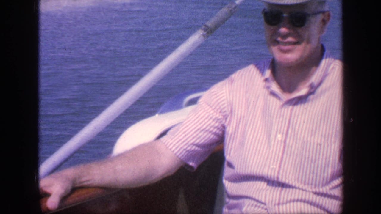 In 1968 Florida home-movie footage, a man in sunglasses, striped shirt, and brimmed hat smiles from a small boat.
