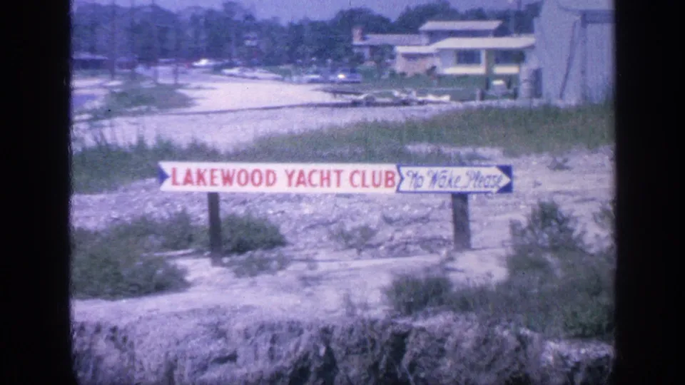 A Lakewood Yacht Club sign appears near a shoreline with water, buildings, grass, and a dock or waterfront area in the background.