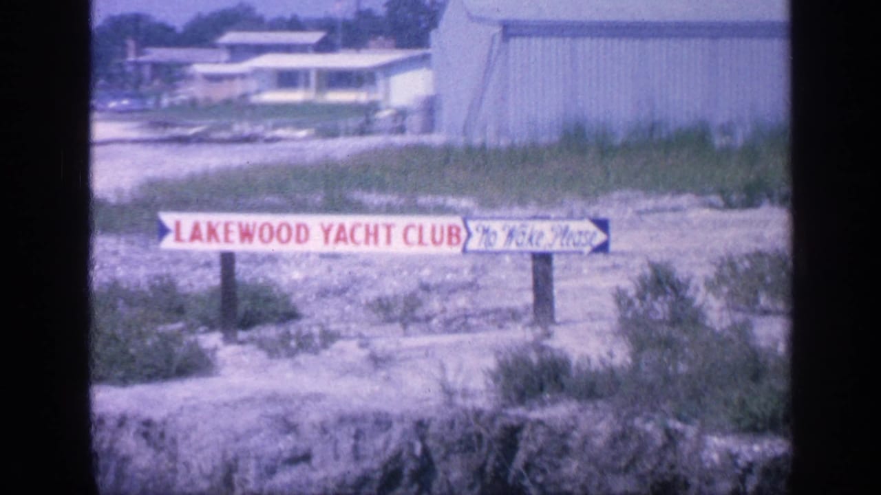 Lakewood Yacht Club sign with No Wake Please text near grass and low marina buildings.