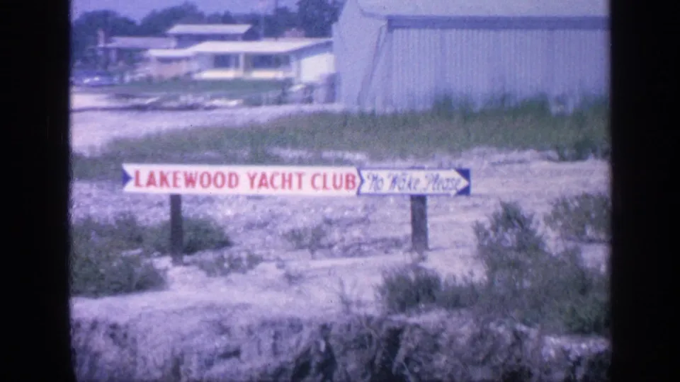 1968 Florida 8mm frame showing a Lakewood Yacht Club sign with No Wake Please text beside a shoreline and modest marina buildings.