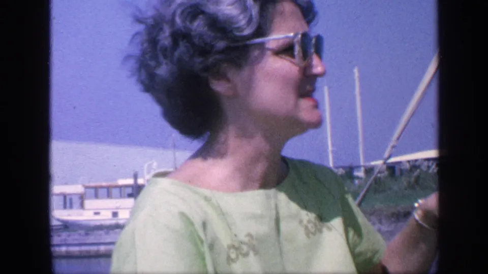 1968 Florida home-movie frame showing a woman in sunglasses and a light green top speaking on a boat with masts and water behind her.