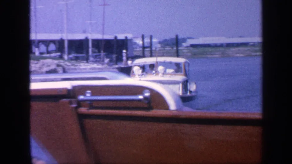 1968 Florida frame showing the stern of a wooden motorboat moving past marina docks, moored boats, water, and a distant shoreline.