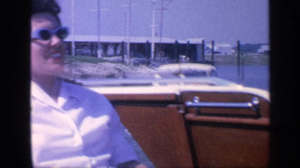 In 1968 Florida home-movie footage, a woman in light blue sunglasses sits in a wooden boat with marina structures behind her.
