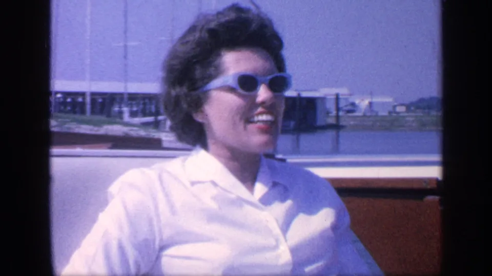 A woman laughs in a wooden boat during a 1968 Florida outing, with calm water and dock pilings behind her.