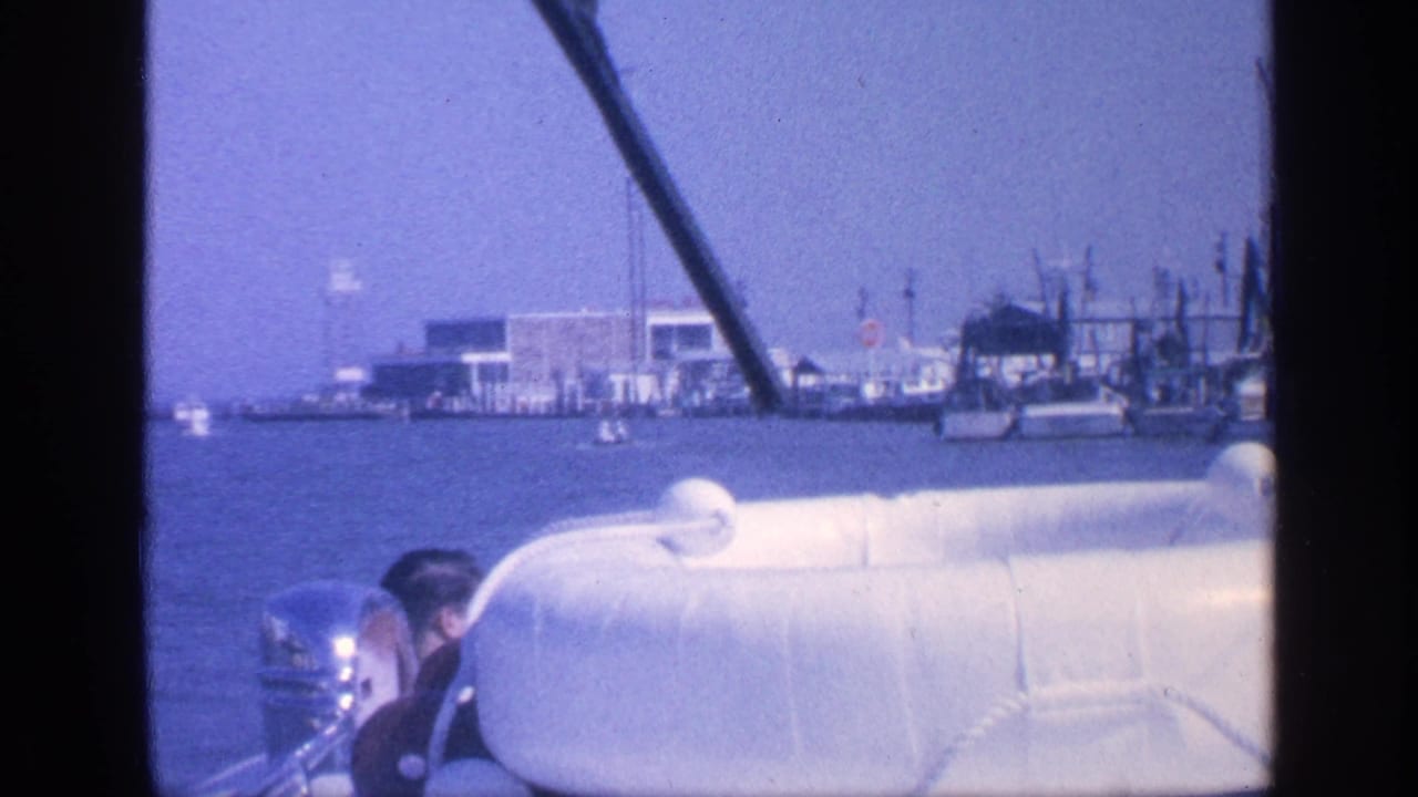 Man partly visible at a boat helm with a white lifeboat in front and port buildings beyond.