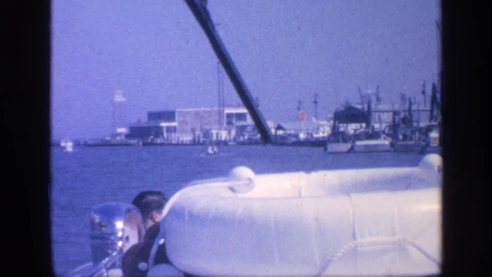 1968 Florida home-movie frame with a partly visible man at the helm, a white lifeboat or seat, outboard motor, and port structures across the water.