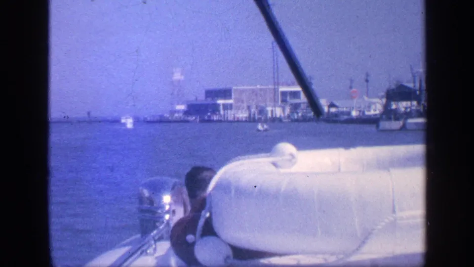 1968 Florida 8mm view from a moving small boat, showing the outboard motor, white foreground fixture, blue water, and distant port buildings.