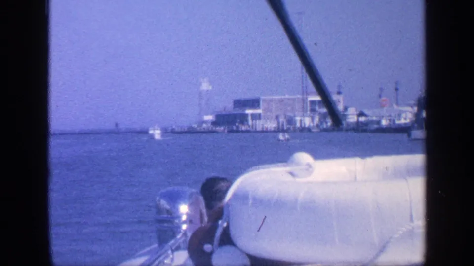1968 Florida port approach frame with the helmsman partly visible behind a white lifeboat or seat, an outboard motor, and industrial waterfront beyond.