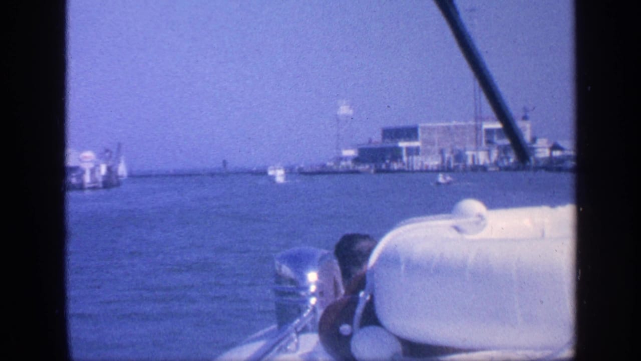 Boat view over calm water with white seat, outboard motor, distant docks, and waterfront buildings.