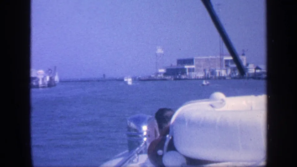 1968 Florida 8mm boat POV with a white padded seat, partial passenger, outboard motor, and distant waterfront structures.