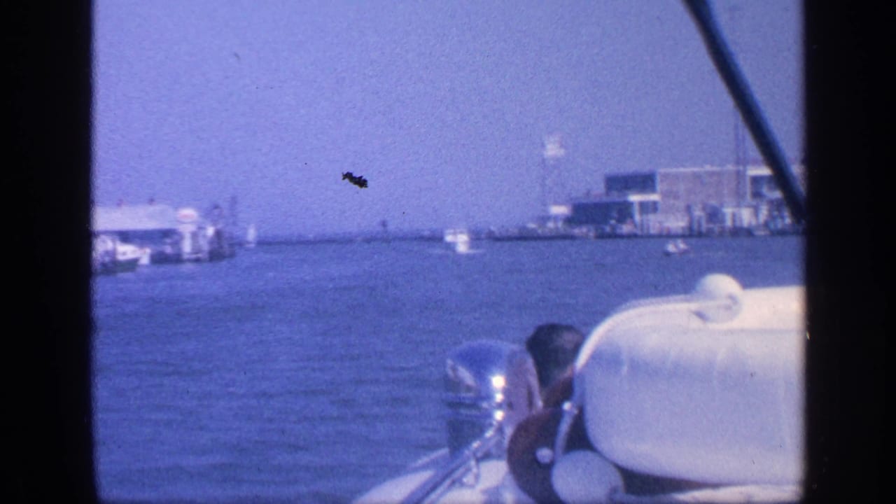 Forward boat view across water with a seated passenger partly visible and buildings along the shore.