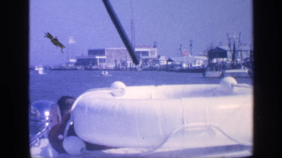 1968 Florida 8mm frame showing a boat cruising past waterfront docks, low buildings, small boats, and a partly visible passenger.