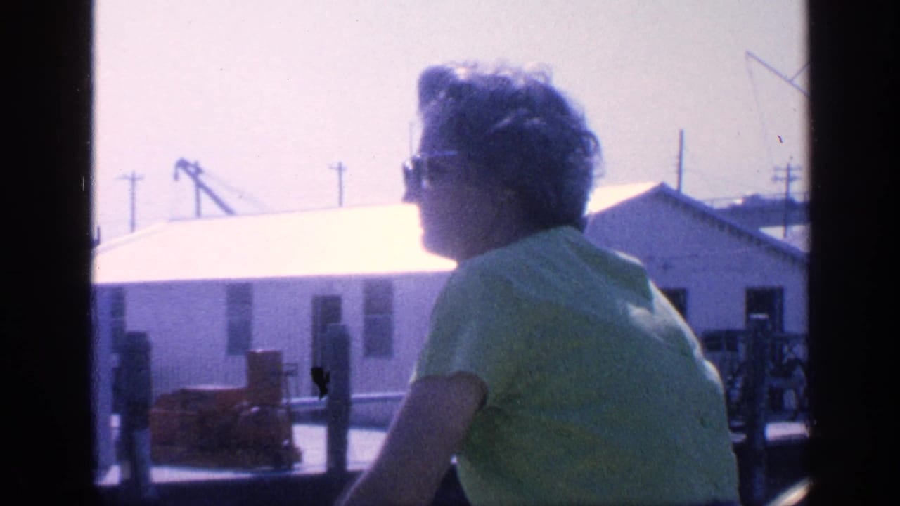 In supplied 1968 Florida 8mm footage, a woman in sunglasses and a green shirt rides past low waterfront buildings.
